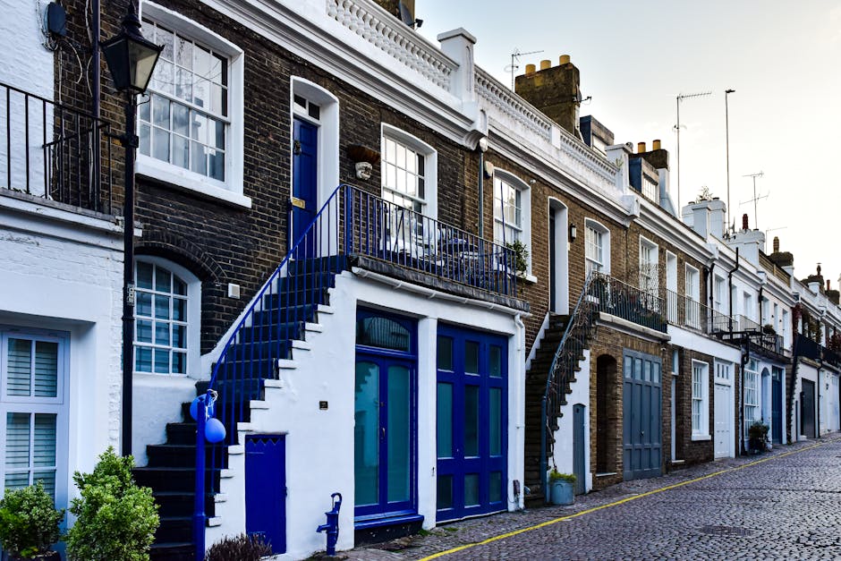A row of terraced houses in Belsize Park featuring brick facades with white decorative trim and large windows. The houses have external staircase entrances with metal railings painted in dark blue, leading up to small first-floor balconies with potted plants and outdoor furniture. The ground floor includes blue-framed doors and windows, some with white shutters. The street is paved with cobblestones, with some greenery in small shrubs near the houses. The scene is well-lit, indicating daytime, and the image depicts a typical residential environment suitable for home relocation and furniture transport services offered by Man With a Van Belsize Park, emphasizing the tight access of these properties.