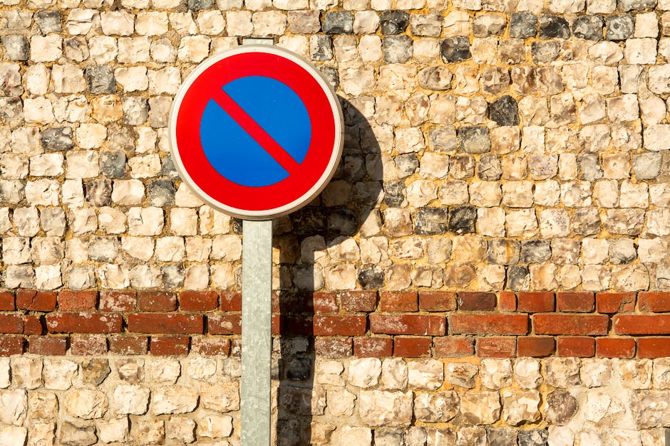 A rectangular traffic sign displaying the message 'No Parking Any Time' in red capital letters on a white background, mounted on a metal pole with multiple holes. The sign is situated outdoors, with g