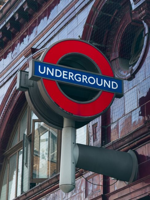 Close-up view of a London Underground station sign with a blue background and white text reading 'UNDERGROUND', mounted on a grey pole with a horizontal grey tube at the bottom. Behind the sign, there is a building with reflective glass windows and dark red-brown tiles, indicating an urban environment. The sign's placement suggests it is part of the infrastructure used during a house removal or home relocation process, which may involve the transportation of furniture and boxes through city transit. Man With a Van Belsize Park provides removals services that include moving belongings along such routes, and the image emphasizes the relevance of urban transport logistics in professional moving operations.
