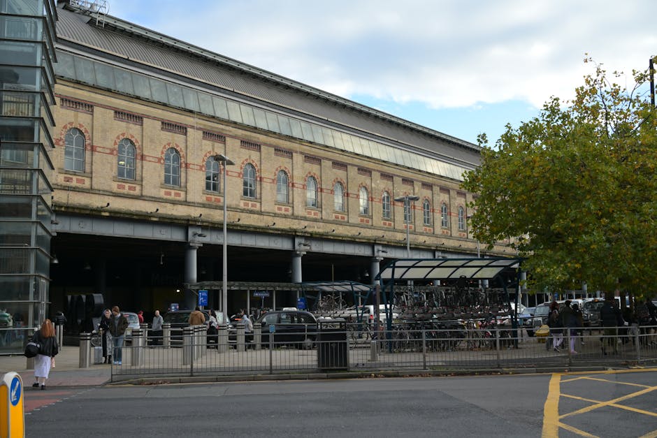 A large brick building with arched windows, situated above a car parking area and bicycle storage, is visible in the image taken during daylight hours. The building's exterior features decorative brickwork with red accents and small rectangular windows along the upper level. The ground level shows a sheltered area with metal columns supporting the structure above, beneath which several cars are parked or waiting. In the foreground, a paved sidewalk runs parallel to the street, separated by a metal railing, with pedestrians walking or standing nearby, some engaged in carrying boxes or bags. To the right, a large tree with green foliage partially obstructs the view of the building, and street lamps and signposts are visible along the pavement. The scene suggests an urban environment with busy foot traffic and vehicle activity, typical of a location involved in house removals and home relocation services, with a focus on loading or unloading items for a move, as managed by [COMPANY_NAME].